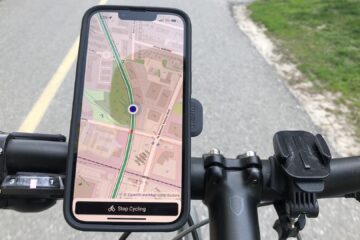A cyclist's view looking down at the handlebars of a bike. There's a phone mounted on them. On the phone's screen, the Cycling Guide app is showing a map. In the background the asphalt trail on which the cyclist is riding can be seen.