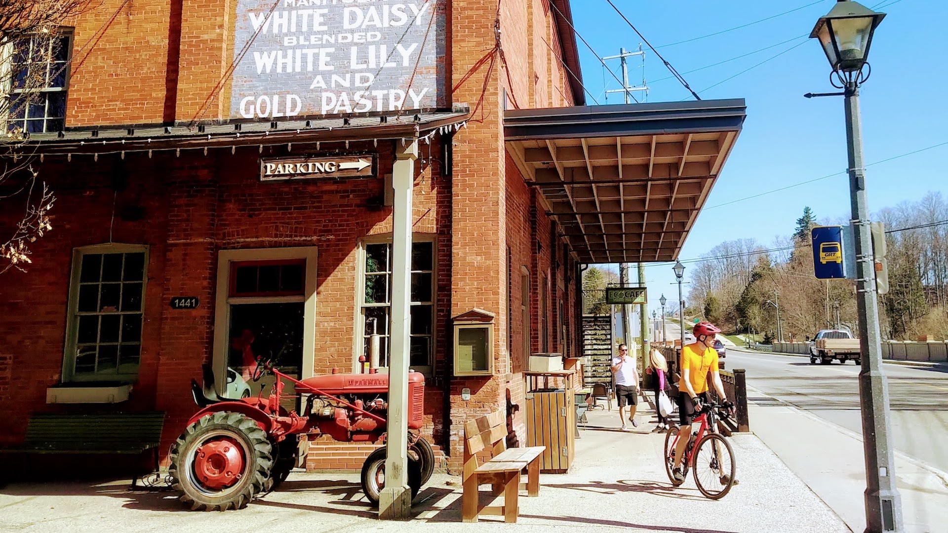 A few of the corner of a red brick building. In the left foreground there's a red farm tractor. In the right foreground there's a person on a bike.