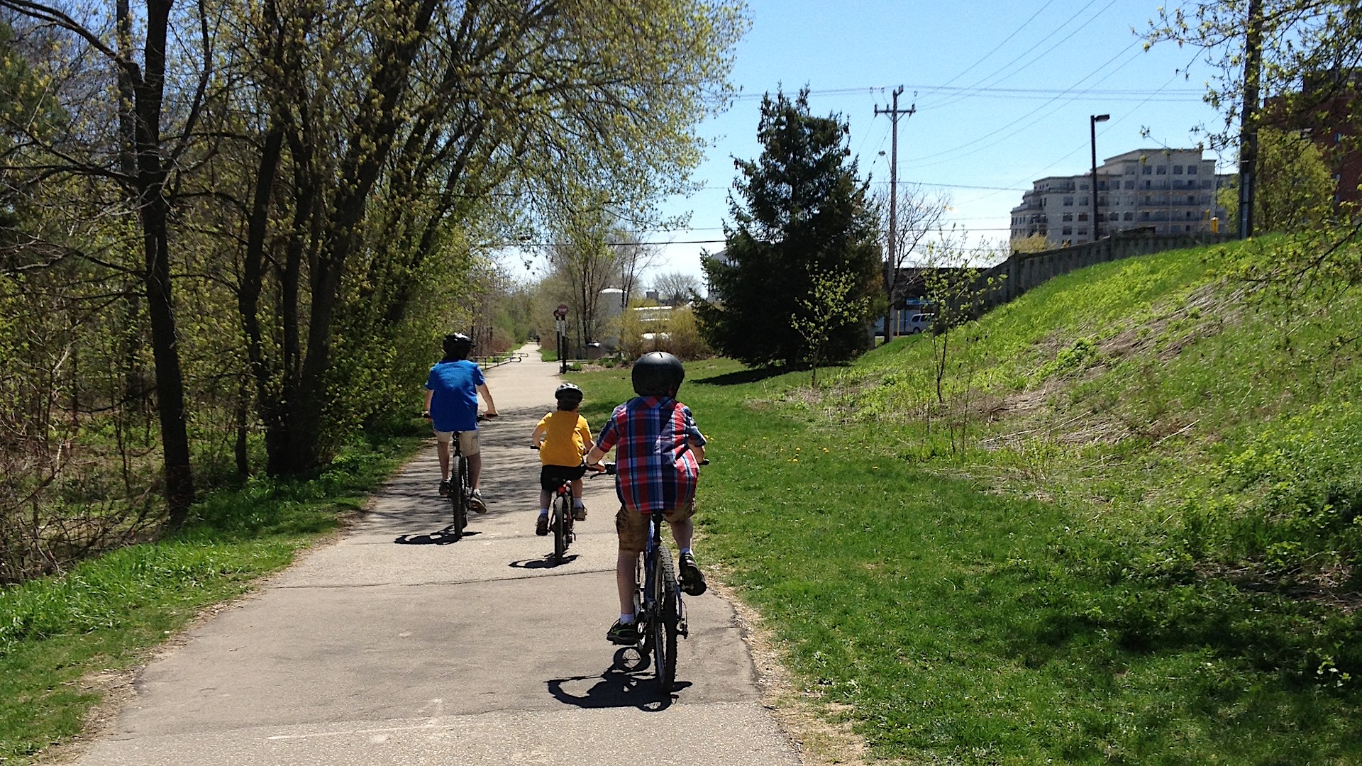 Three children on bikes are cycling away from the viewer on an asphalt multi-use path. Grass and trees are visible on either side of the path.