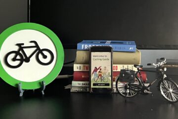 A desktop has some cycling-related artifacts: the Cycling Guide app icon, 6 cycling-related books, a phone with the Cycling Guide initial screen, and a model bicycle.