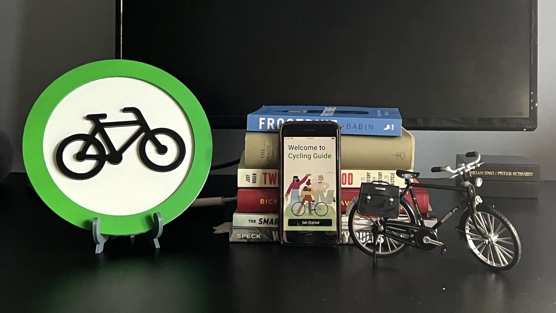 A desktop has some cycling-related artifacts: the Cycling Guide app icon, 6 cycling-related books, a phone with the Cycling Guide initial screen, and a model bicycle.