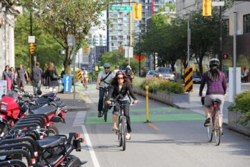 A view of an urban street. Cyclists approach the viewer in a protected bike lane. There are people walking on a sidewalk on the left, and cars moving in their own lanes on the right.