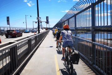 People on bikes are riding on a protected multi-use path that's on a bridge. On the left, on the other side a strong barrier, there are lanes of car traffic. It's a sunny day.