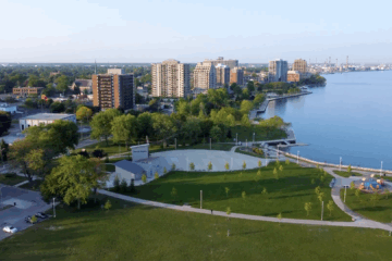 An aerial view of the waterfront in Sarnia. In the foreground there's green space and paths. In the middle ground there are leafy trees and apartments. On the right, the blue water of Lake Huron is visible. The sky is clear.