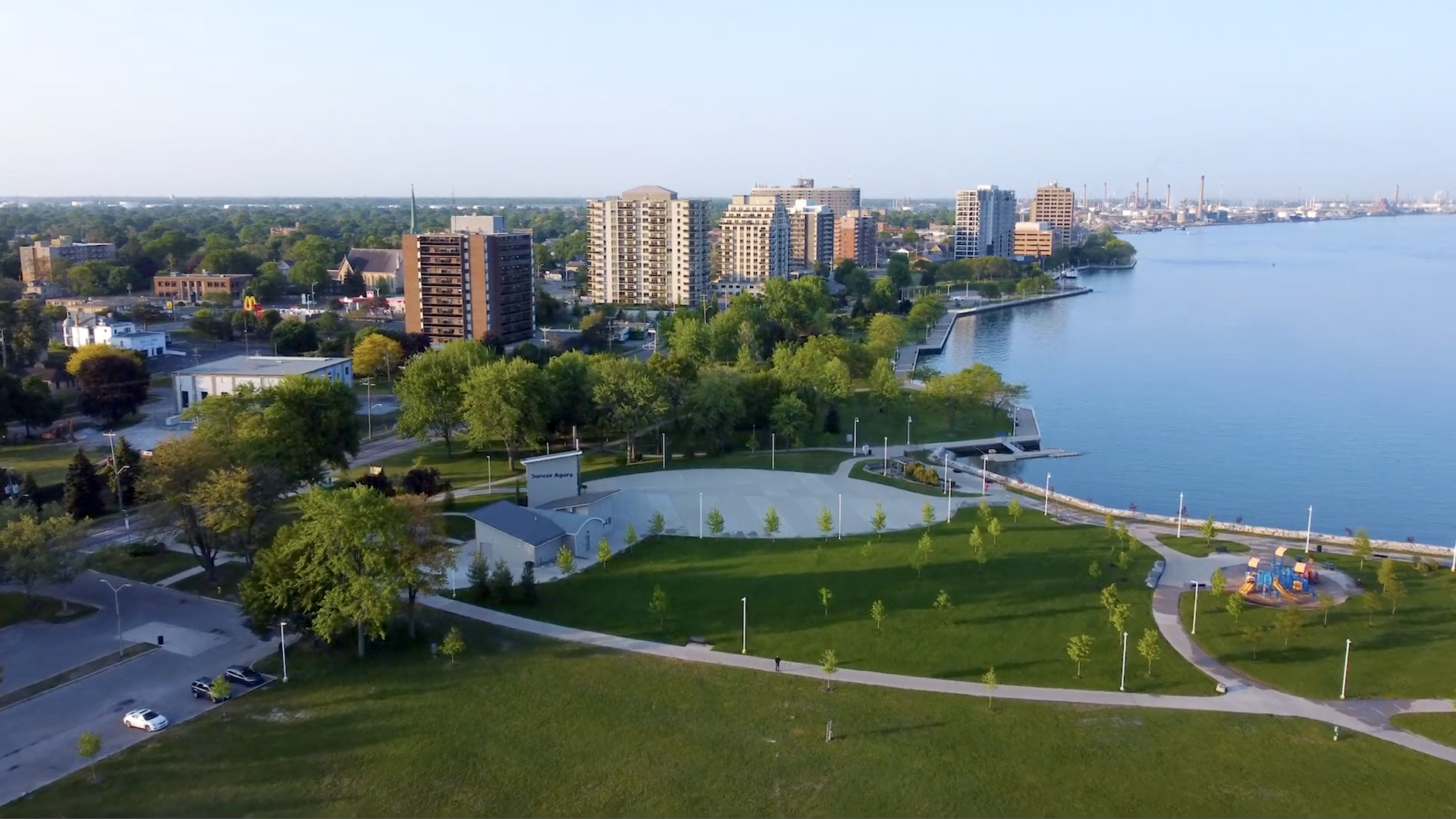 An aerial view of the waterfront in Sarnia. In the foreground there's green space and paths. In the middle ground there are leafy trees and apartments. On the right, the blue water of Lake Huron is visible. The sky is clear.