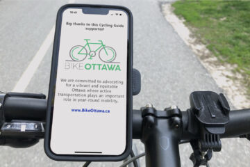 A cyclist's view looking down at the handlebars of a bike. There's a phone mounted on them. On the phone's screen, the Cycling Guide app is showing a sponsor screen for Bike Ottawa. In the background the asphalt trail on which the cyclist is riding can be seen.