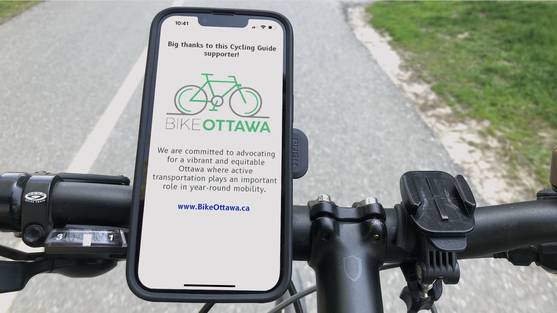 A cyclist's view looking down at the handlebars of a bike. There's a phone mounted on them. On the phone's screen, the Cycling Guide app is showing a sponsor screen for Bike Ottawa. In the background the asphalt trail on which the cyclist is riding can be seen.