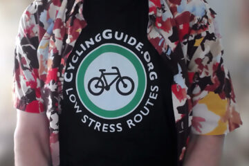 A view of the upper torso of someone. They're wearing a colourful short-sleeved buttun up shirt that's open, revealing a black t-shirt with a white and green Cycling Guide log, with text that reads "CyclingGuide.org Low Stress Routes"