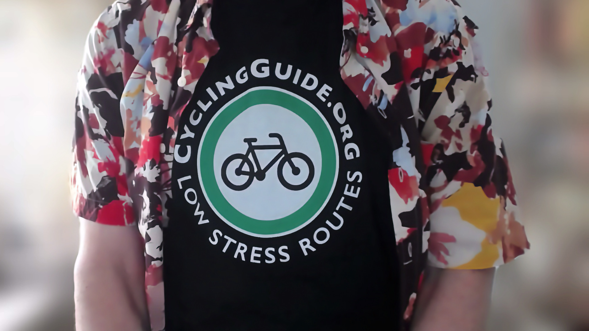 A view of the upper torso of someone. They're wearing a colourful short-sleeved buttun up shirt that's open, revealing a black t-shirt with a white and green Cycling Guide log, with text that reads "CyclingGuide.org Low Stress Routes"