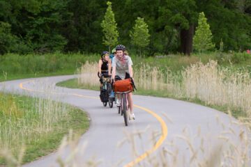 A curvy multi-use path recedes into distant trees. Three people on bikes are approach, and they all look happy!