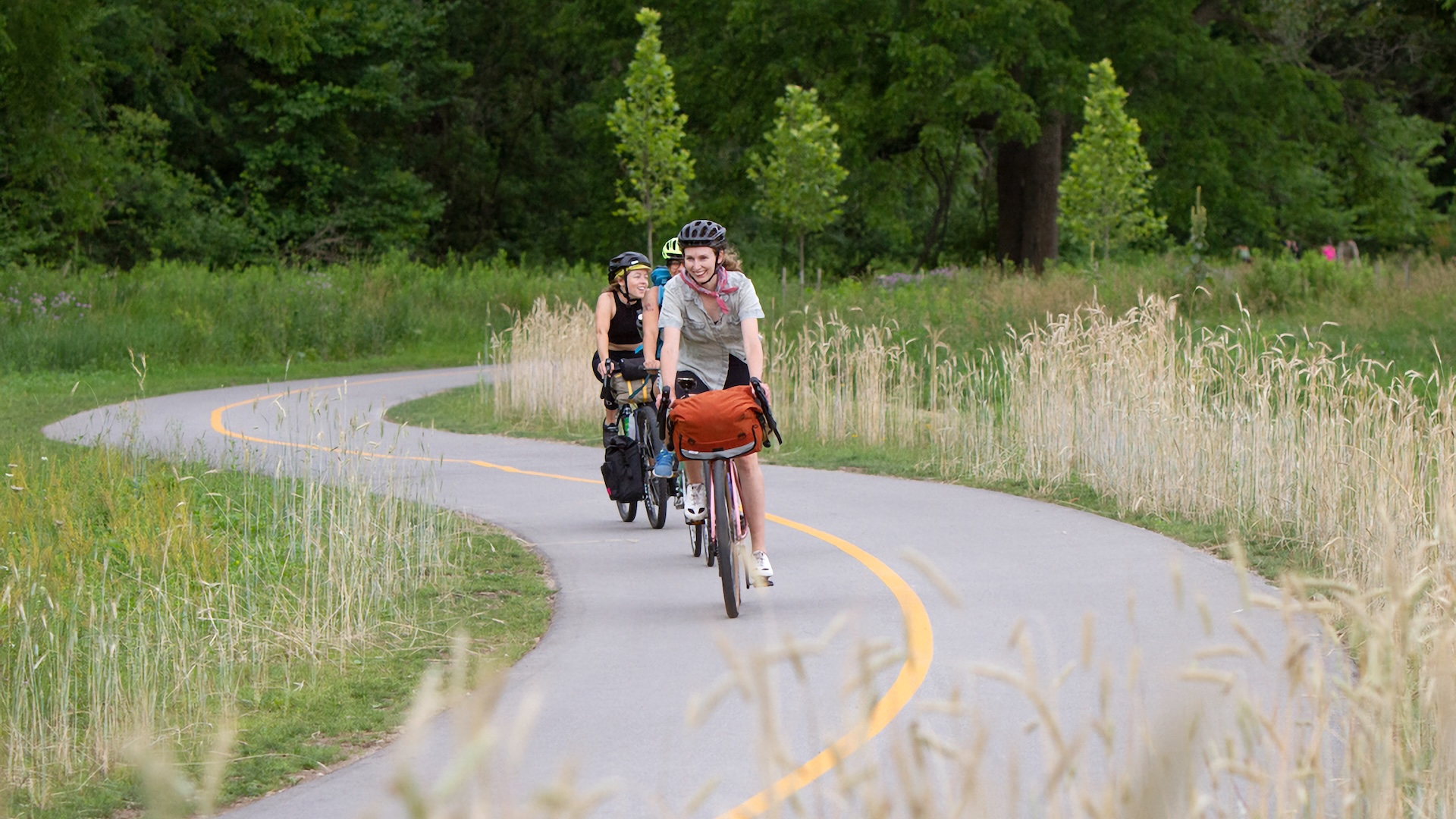 A curvy multi-use path recedes into distant trees. Three people on bikes are approach, and they all look happy!