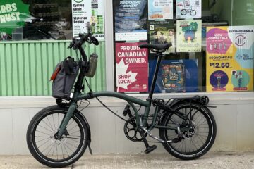 In the foreground, there’s a Brompton bicycle leaning against a storefront. Behind the Brompton in the store window, there are multiple posters, including one for Cycling Guide.