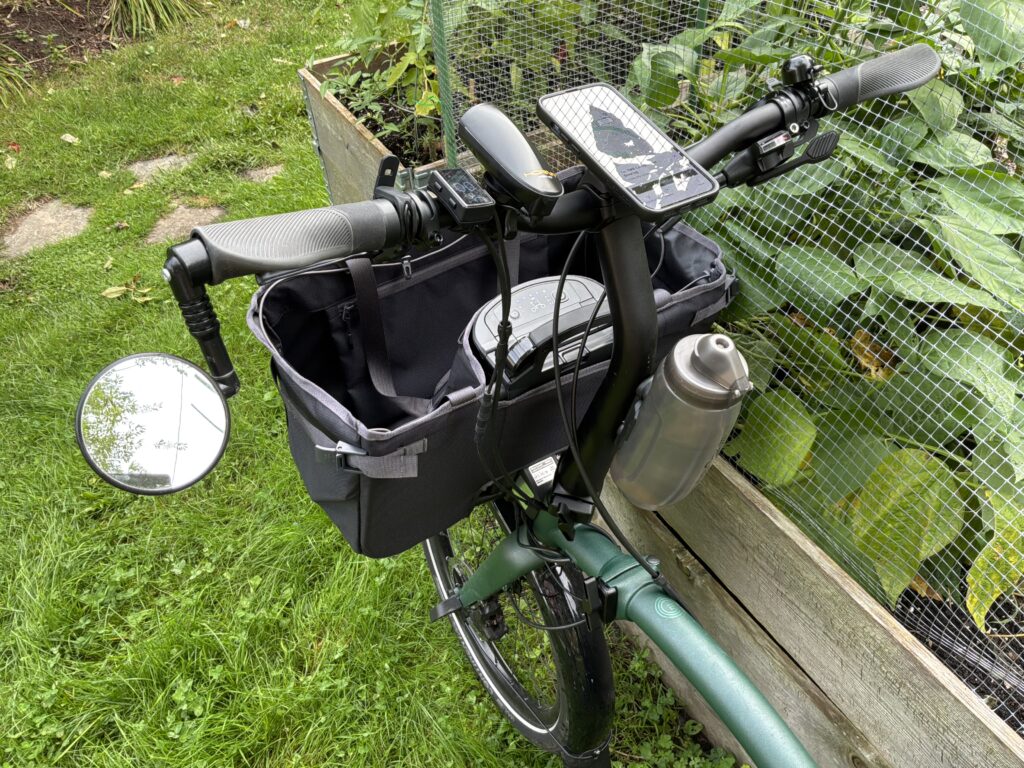 An overhead view of the handlebars of a bike. From left to right theres a mirror, a button for a horn, a bike controller, a horn, a phone on a mount, and a bell. There's a waterbottle mounted on the tube leading to the handlebars. There's a large bag hanging off the front.