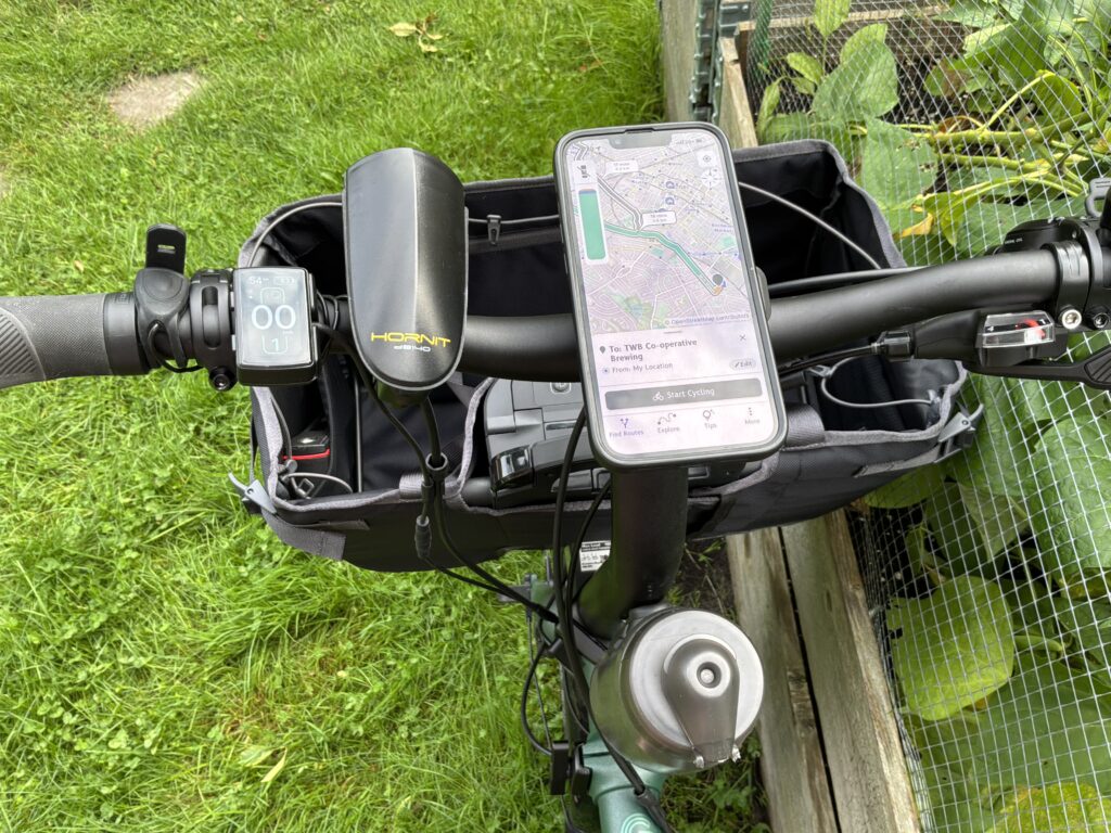 An overhead view of the handlebars of a bike. From left to right there's a button for a horn, a bike controller, a horn, a phone on a mount with a map on its screen, and a bell. There's a waterbottle mounted on the tube leading to the handlebars. There's a large bag hanging off the front.