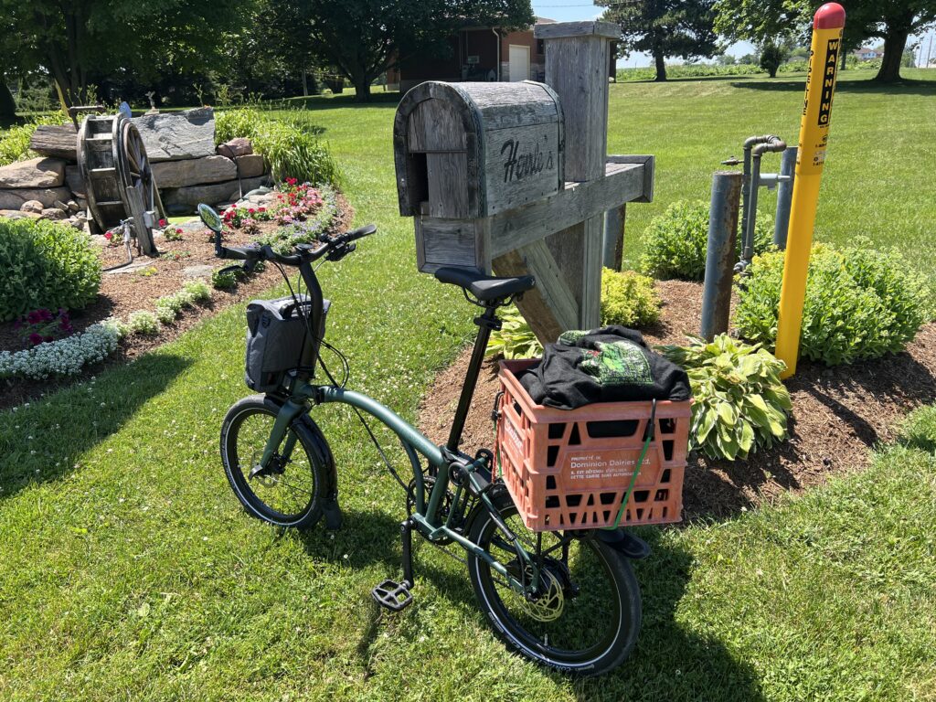 In the foreground, there’s a Brompton bicycle leaning against a rural mailbox. There’s a red milk crate mounted on the back of the bike, such that the sides are at a 45º angle to the rack it's on. There are flowers and other plants in two gardens, and trees in the background. It's a sunny day.