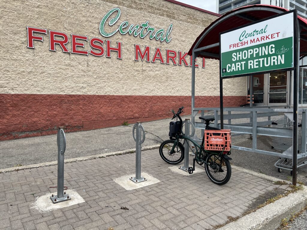 A view of a bike parking area, a Brompton locked to a post-and-ring style parking spot. A large crate is mounted on the rerar rack of the bike. In the background there’s the wall of a building. On the wall the words “Central Fresh Market” are prominent.