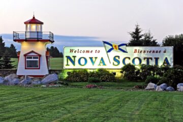 A lighthouse on a green lawn To the right there a large sign that reads "Welcome To Nova Scotia"
