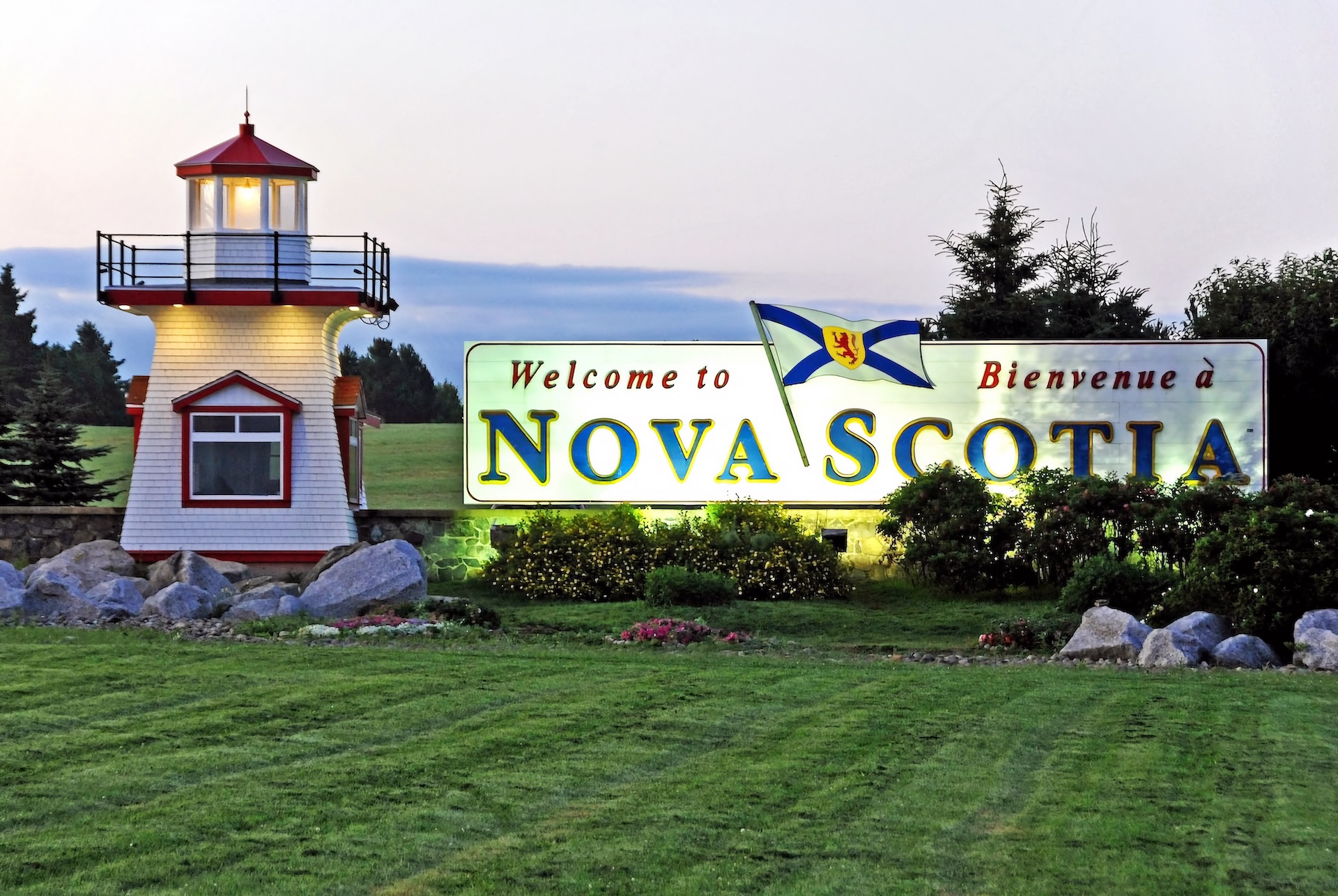 A lighthouse on a green lawn To the right there a large sign that reads "Welcome To Nova Scotia"